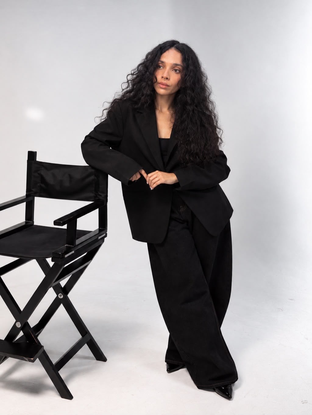 Paola Zuleta, founder of KEWA, in a studio portrait. She leans on a black director's chair in a black blazer and wide-leg trousers, looking away from the camera, against a light grey backdrop.